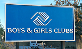 Blue sign with white text and logo for Boys & Girls Clubs, featuring two interlocking hands. A tree and parked car are visible in the background.