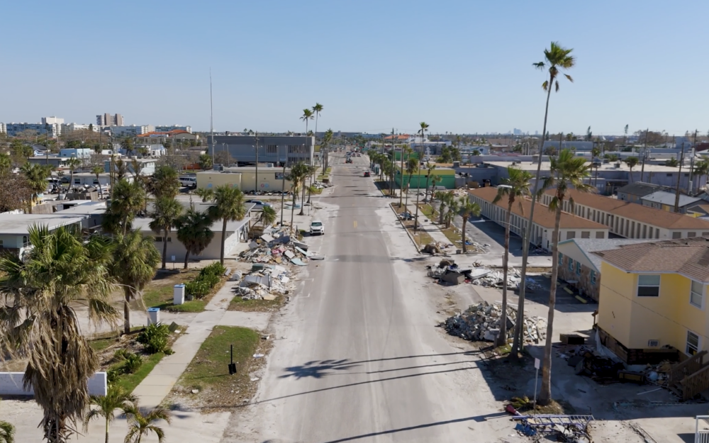 A deserted street lined with palm trees and buildings shows scattered debris and piles of rubbish along the sidewalks under a clear blue sky.