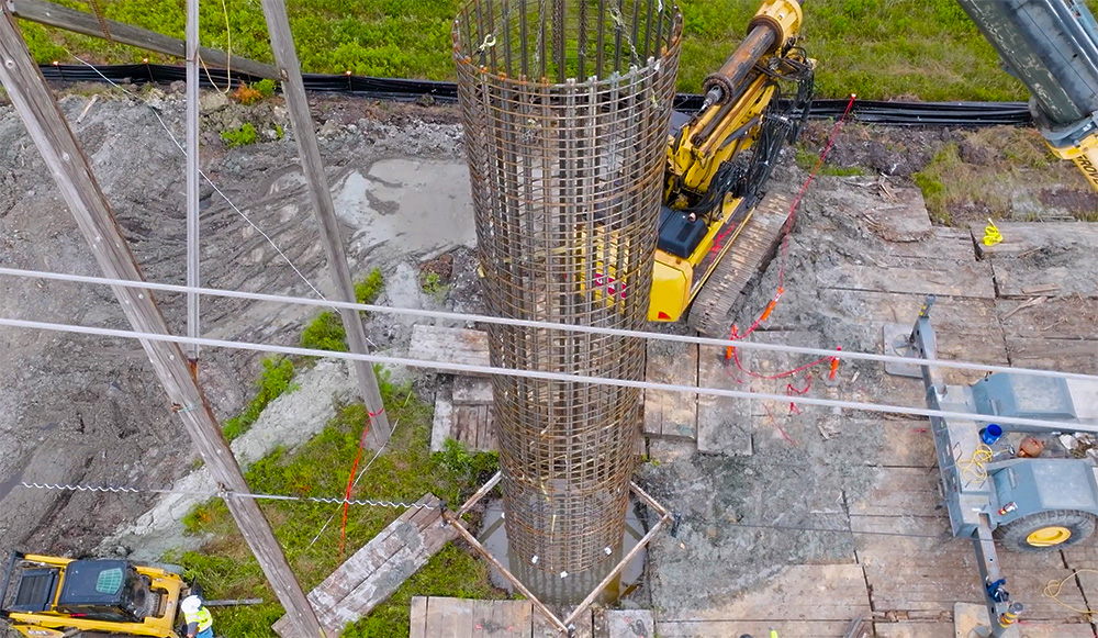 Aerial view of a construction site with a large circular metal rebar structure in the center. Surrounding it are excavators and workers on a muddy, wooden platform.