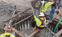 Two construction workers in safety harnesses and hard hats guide a large cylindrical concrete form into a rebar-lined hole filled with wet concrete. A crane hook suspends the form while the workers balance on wooden supports above the foundation area.