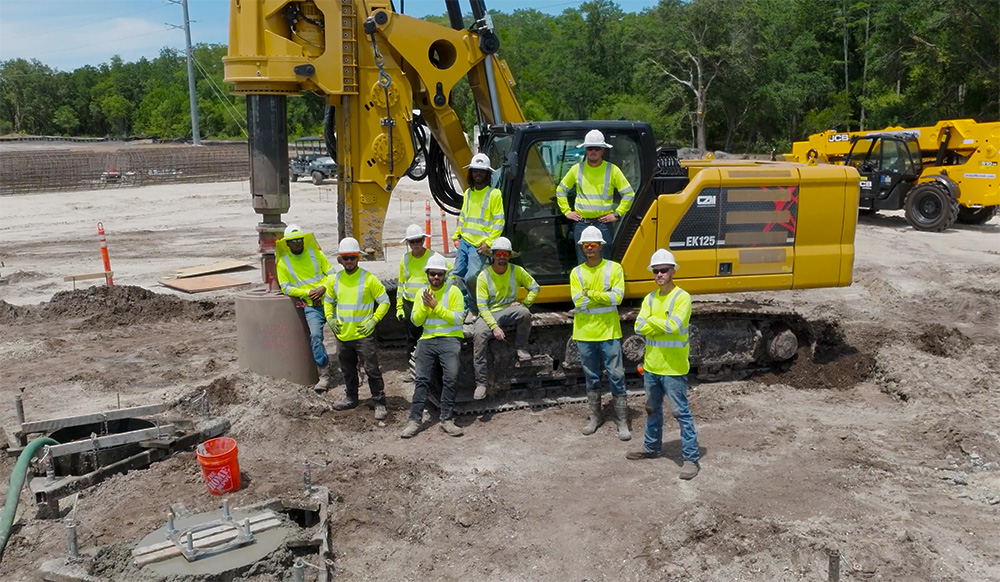 A group of construction workers wearing high-visibility neon yellow shirts and white hard hats stand in front of a large yellow drilling rig at an active construction site. The crew poses on and around the tracked machinery, with dirt ground and partially formed concrete structures visible in the foreground.