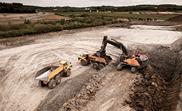 Aerial view of a construction site with two large yellow dump trucks and an excavator at work, surrounded by expansive earth and greenery under a cloudy sky.