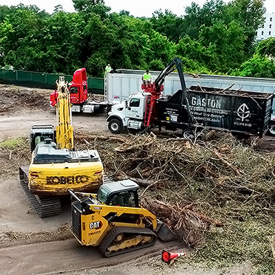 A construction site with heavy machinery, including a yellow Kobelco excavator and a CAT loader, moving debris into two large Gaston trucks. Piles of branches cover the ground; trees and a building are visible in the background.
