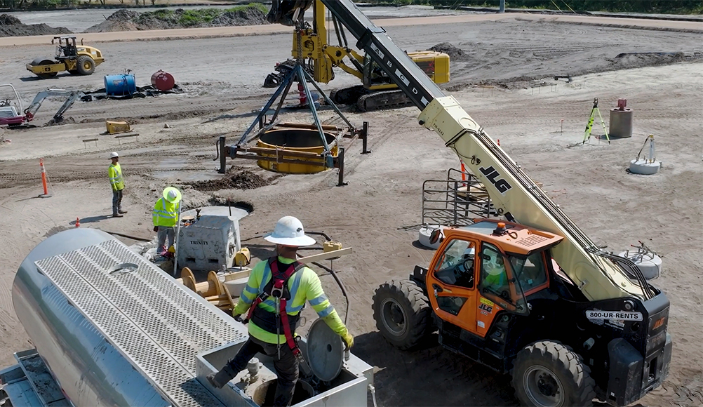 Construction site with a yellow crane lifting heavy equipment, a worker in safety gear guiding from below. A JLG telehandler is nearby, conveying teamwork and precision.