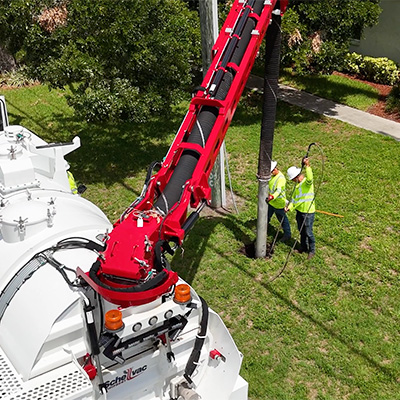 Two workers in safety vests and helmets operate a large white vacuum truck with a red hydraulic arm, cleaning out a utility hole in a grassy area near a sidewalk and residential building.