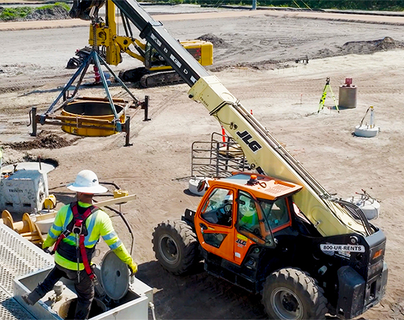 Construction site with a yellow crane lifting heavy equipment, a worker in safety gear guiding from below. A JLG telehandler is nearby, conveying teamwork and precision.