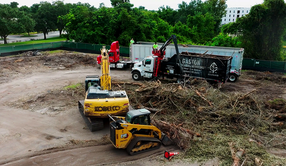 A construction site with heavy machinery, including a yellow Kobelco excavator and a CAT loader, moving debris into two large Gaston trucks. Piles of branches cover the ground; trees and a building are visible in the background.