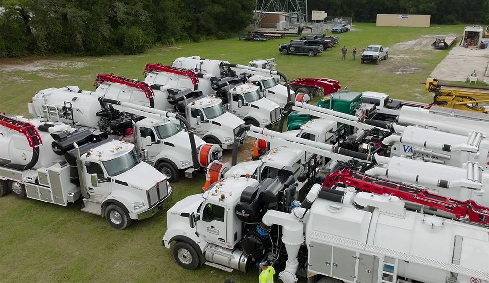 A fleet of white utility vac trucks with red accents is parked in rows on a grassy lot, surrounded by trees. Several people and vehicles are visible nearby. A logo in the bottom right reads Trinity Co.