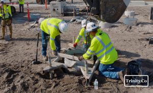 Three construction workers in yellow safety jackets and helmets position a concrete structure at a sandy worksite. Equipment and tools are visible, and a Trinity Co logo appears in the bottom right corner.